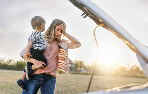 woman holding a child and her head looking at the hood of her broke down car