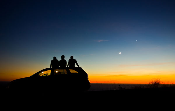 three people sitting on top of a car looking up at the sky in the dusk